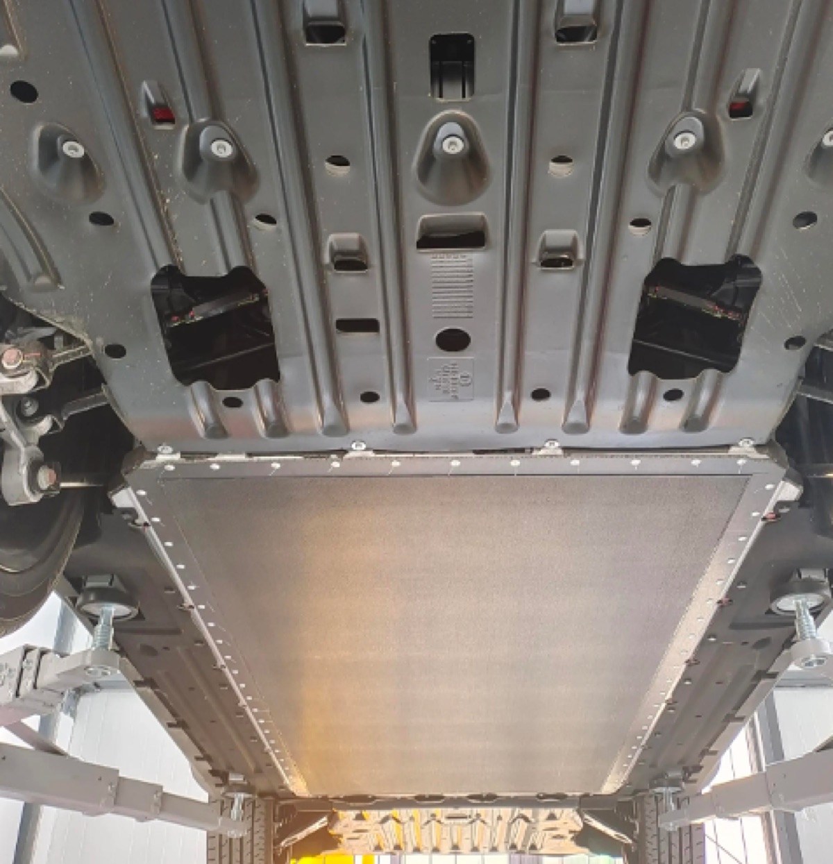 The underbody of an electric vehicle on a lift, showing a large flat battery cover panel